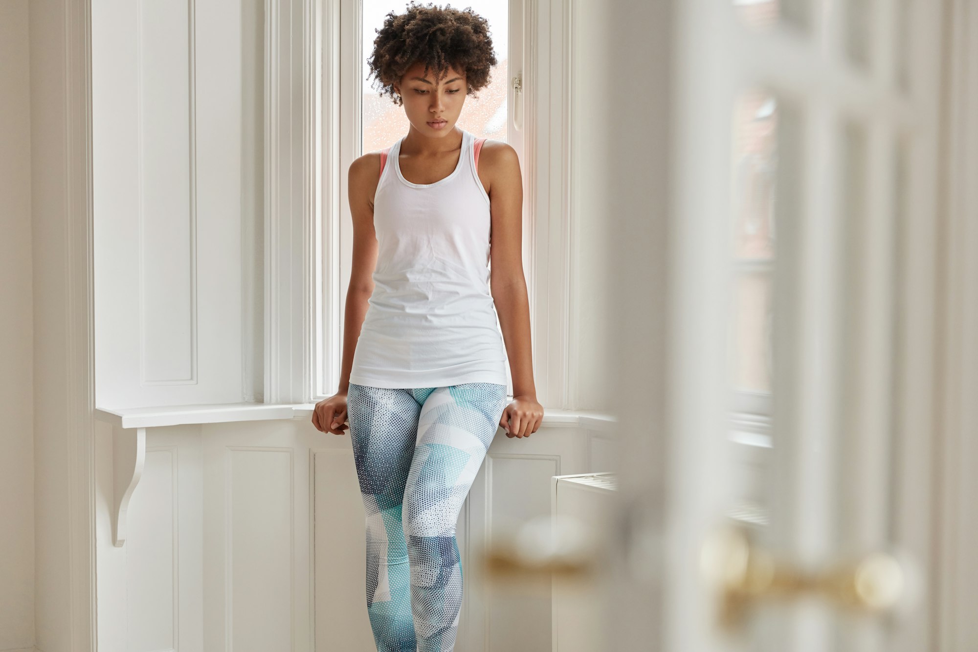 Horizontal shot of beautiful thoughtful dark skinned girl dressed in casual white t shirt and leggin
